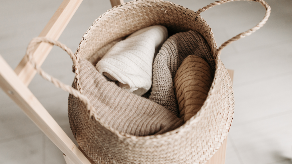 A woven basket containing three folded, neutral-toned knit sweaters sits on a wooden chair, ready for your next closet cleanout.